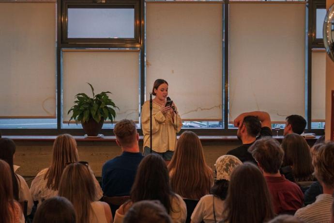 A speaker stands on stage with a microphone, engaging an attentive audience in a cozy, well-lit venue.