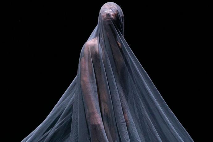 A figure stands shrouded in a sheer gray fabric, partially obscuring their body and face, against a dark background.