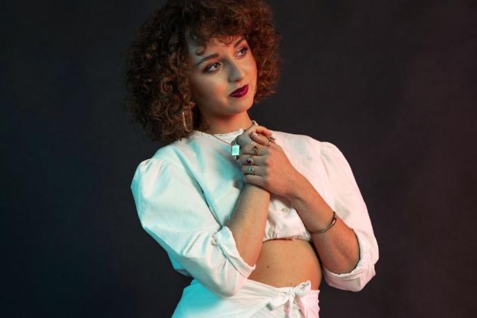 A person with curly hair poses gracefully in a stylish white outfit, showcasing intricate jewelry against a dark, moody background.