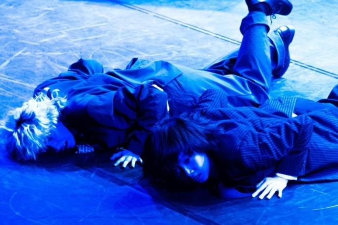 Two performers in formal attire lie on a dark stage, illuminated by blue lighting.
