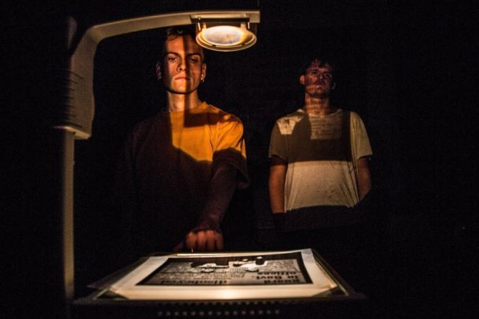 Two figures stand in dim lighting, one casting a shadow on a lightbox displaying an illustrated document.