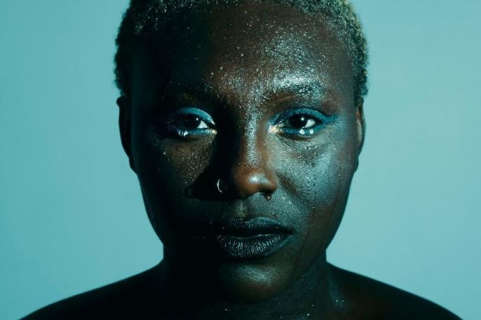A person with black skin wearing blue eyeshadow, with prominent cheekbones, captured in a portrait shot indoors.