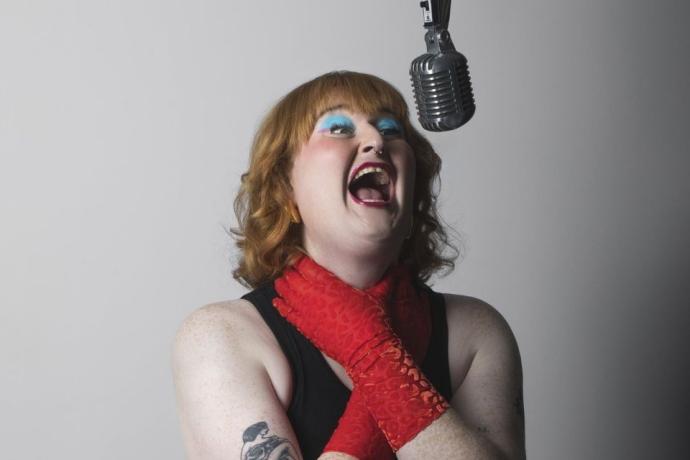 A woman singing into a hanging microphone, wearing lipstick and fashionable clothing, with her shoulder and elbow visible as she holds her neck