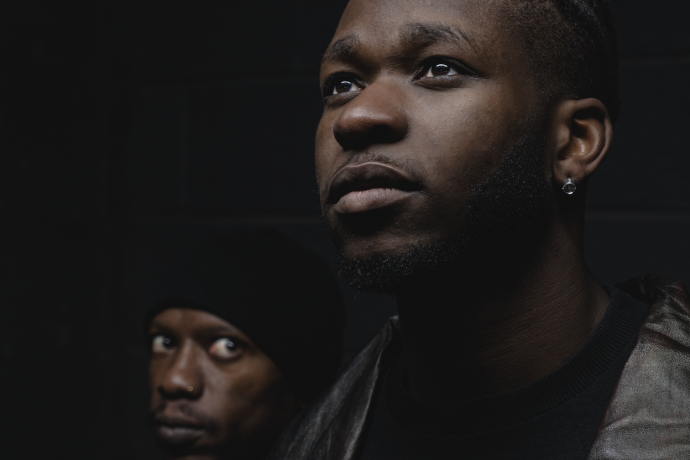 Two men standing next to each other against a black background, with one man staring at the other intensively 