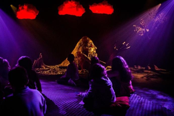 A group of children sit cross-legged on cushions in a dimly lit theatre space, watching an abstract, nature-inspired performance. The stage features a glowing central figure made of textured materials, resembling a mountain or creature, with dramatic purple and red lighting. Red clouds hang above, and a tree stands to the right, creating a dreamlike, immersive atmosphere.