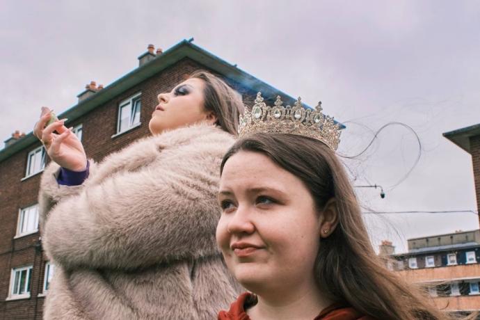 A young girl wearing a tiara stands in the foreground, while a woman in a fur coat vapes in the background, with residential buildings behind them under a cloudy sky.
