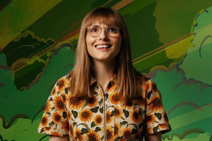A smiling woman with long light brown hair and glasses sits against a vibrant, illustrated green background of stylised trees, clouds, and a bus. She wears a sunflower-print zip-up dress and appears cheerful and relaxed in the centre of the frame.