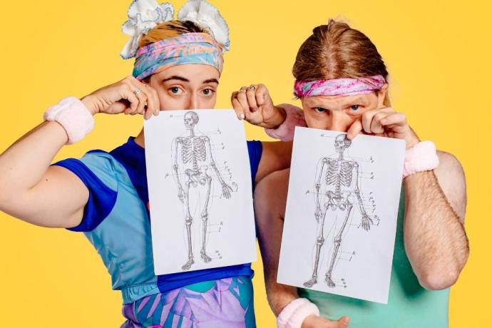 Colourful promotional image of two performers in retro fitness costumes holding anatomical skeleton diagrams. Quirky, inclusive theatre show about disability, bodies, and movement. Bold yellow background, 80s-inspired fashion, and comedy performance imagery.