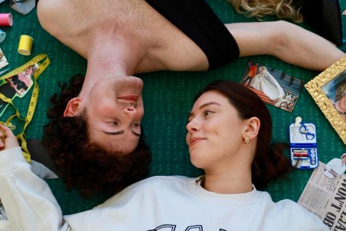 A man and woman relax on the floor surrounded by random objects, looking at each other's eyes.