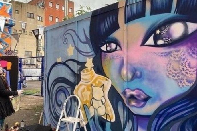 A colorful mural featuring a woman with blue hair holding a yellow bottle, surrounded by vibrant street art elements