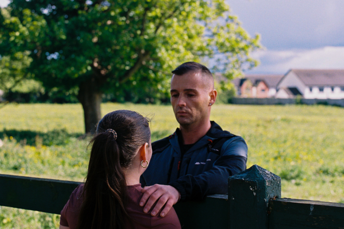 Two people standing by a wooden fence outdoors, with one person placing a hand on the other's shoulder against a backdrop of a grassy field and a large tree.
