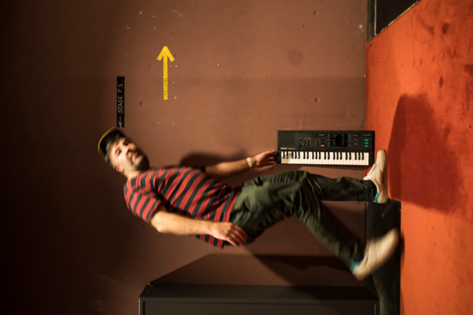 A sideways-oriented photograph of a man in a striped shirt appearing to float horizontally next to a wall keyboard backstage.