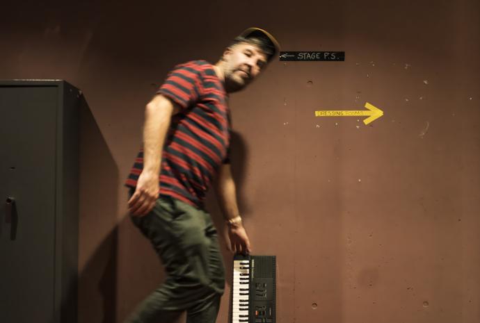 A man in a striped shirt and baseball cap holding a small electronic keyboard vertically against a brown backstage wall with hand-painted arrows for "STAGE P.S." and "DRESSING ROOMS."