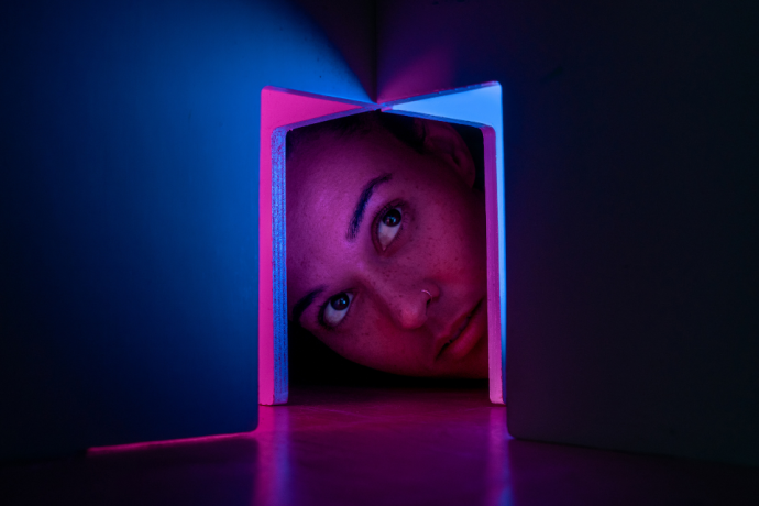 A close-up of a person’s face partly framed inside a small box, lit with pink and blue light in a dark space.
