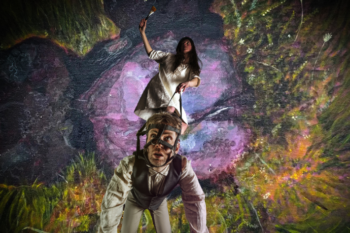 A dancer in a red dress and dark jacket appears doubled and mirrored against a misty landscape. 