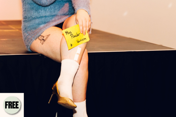 A seated performer in a blue dress and gold heel holds a yellow note reading ‘Miss Powder Vacuum’ on their leg. Visible symbol: Free.