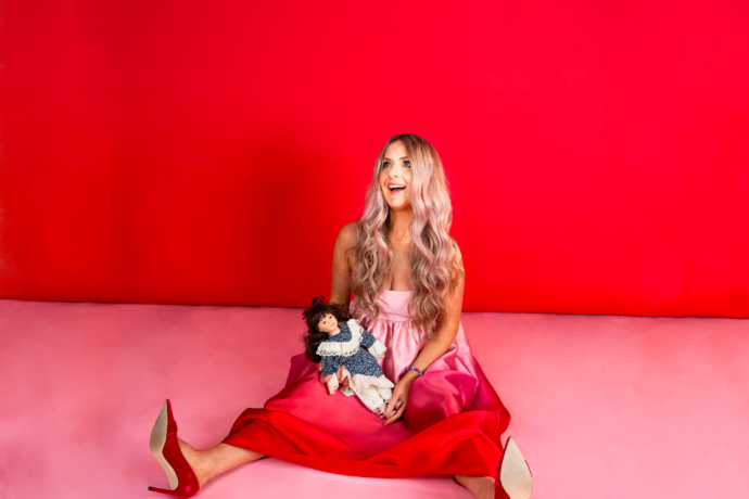 A woman in a pink dress sits against a red and pink studio background, holding a doll in her lap.