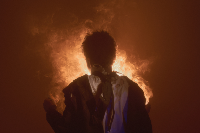 A silhouetted performer stands with bright orange smoke or fire-like light glowing behind them.