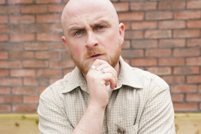A close-up portrait of a bald, bearded man with his hand on his chin in a thinking pose against a brick wall.
