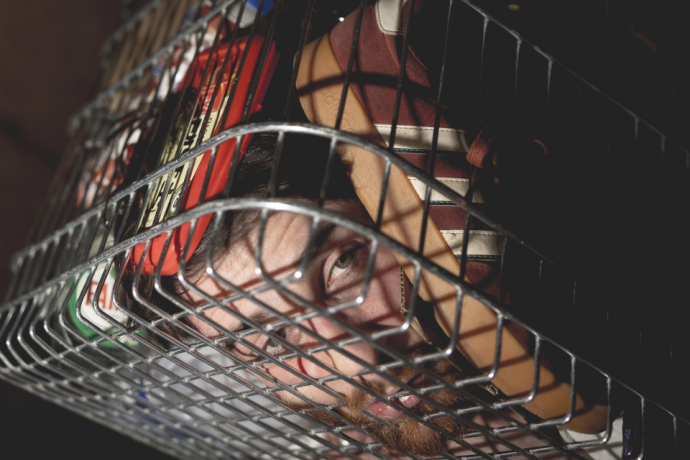 A close-up of a person’s face partly hidden behind the metal bars and shadows of a shopping trolley.