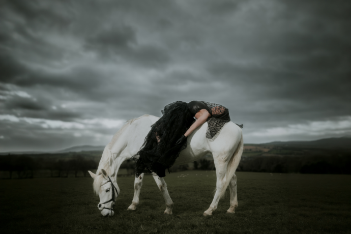 A woman draped over a white horse in a dark rural landscape under heavy grey clouds.