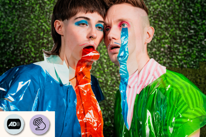 Two performers in shiny blue and green outfits pose close together with bright coloured streamers in their mouths. Visible access symbol: Touch Tours and Audio Description.