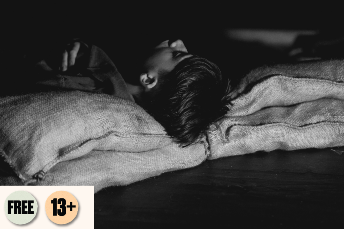 A black and white close-up photo of an child lying down with their head resting on coarse burlap sacks in dim lighting.Visible access symbol: Free and 13+.