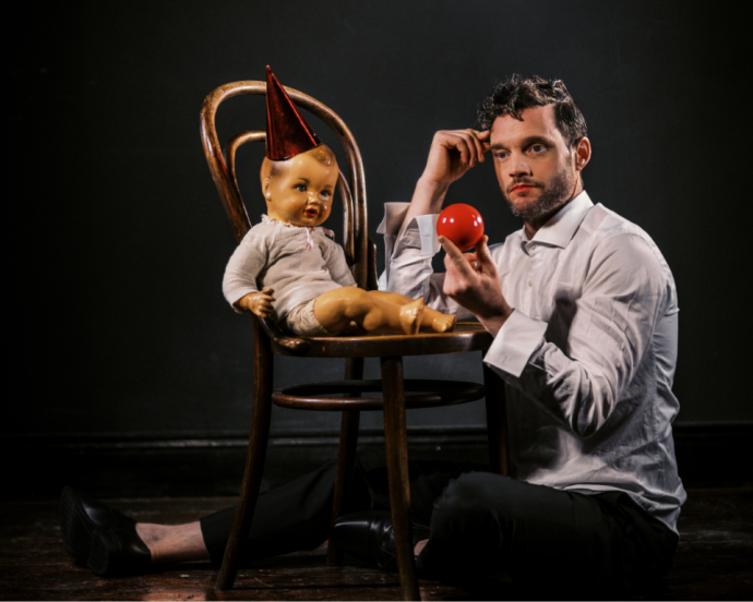 A person in a white shirt sits on the floor holding a red nose, looking thoughtfully at a vintage doll wearing a party hat on a wooden chair.