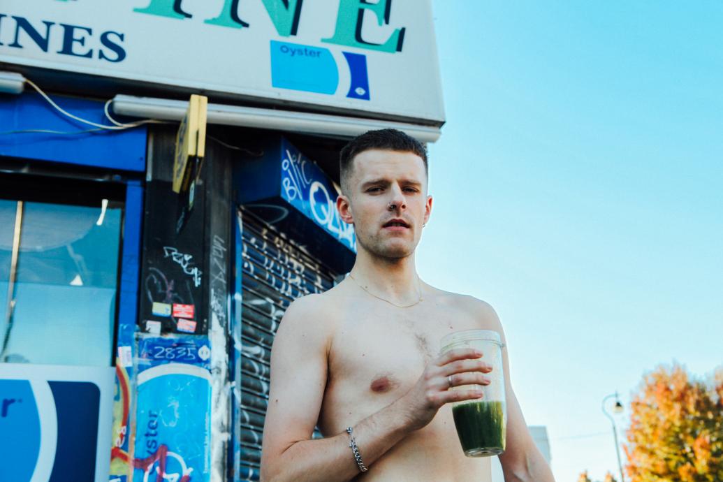 A topless man stands outside a shop during the day time holding a green smoothie