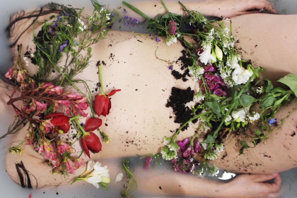 A woman's body floats in a full bath, her body covered in flowers and soil
