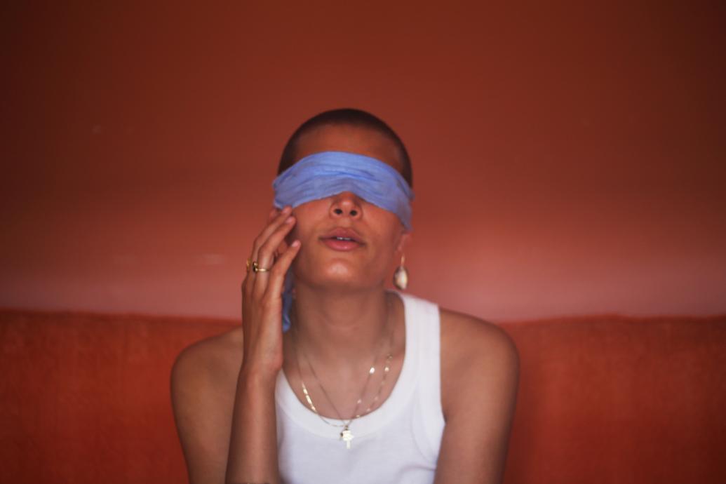 A woman in a white top sits on a red sofa in a red room with a blue blindfold over her eyes and a hand to her face