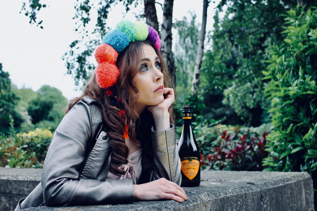 Modern Juliette in flower crown and biker jacket looking out wistfully from a balcony window, with a Buckfast beside her.