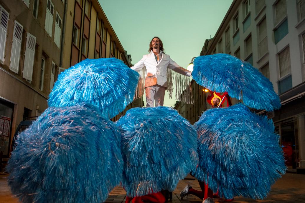 A man dressed in a white suit with fringed sleeves stands on top of a group of umbrellas covered in blue tinsel held by people wearing flame headdresses