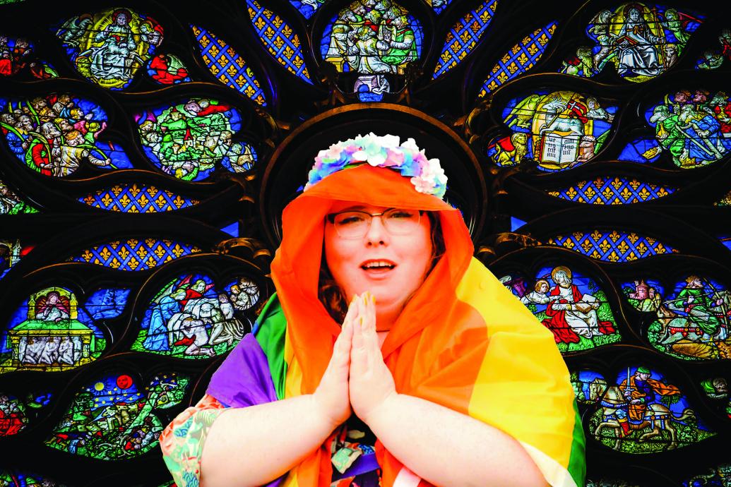 A woman poses against a stained glass window like the Virgin Mary, her hands held in prayer, her veil is a Pride flag and she wears a flower crown