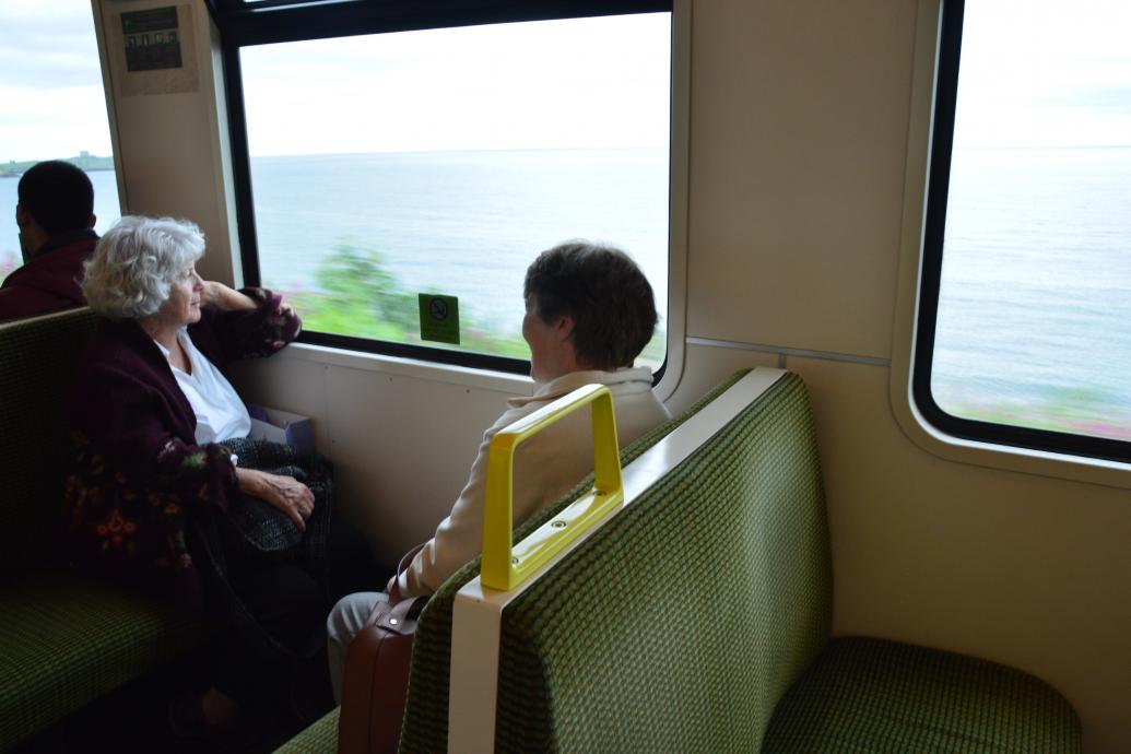 Two women sit facing each other on the dart, the one facing camera looks out the window at the sea passing by