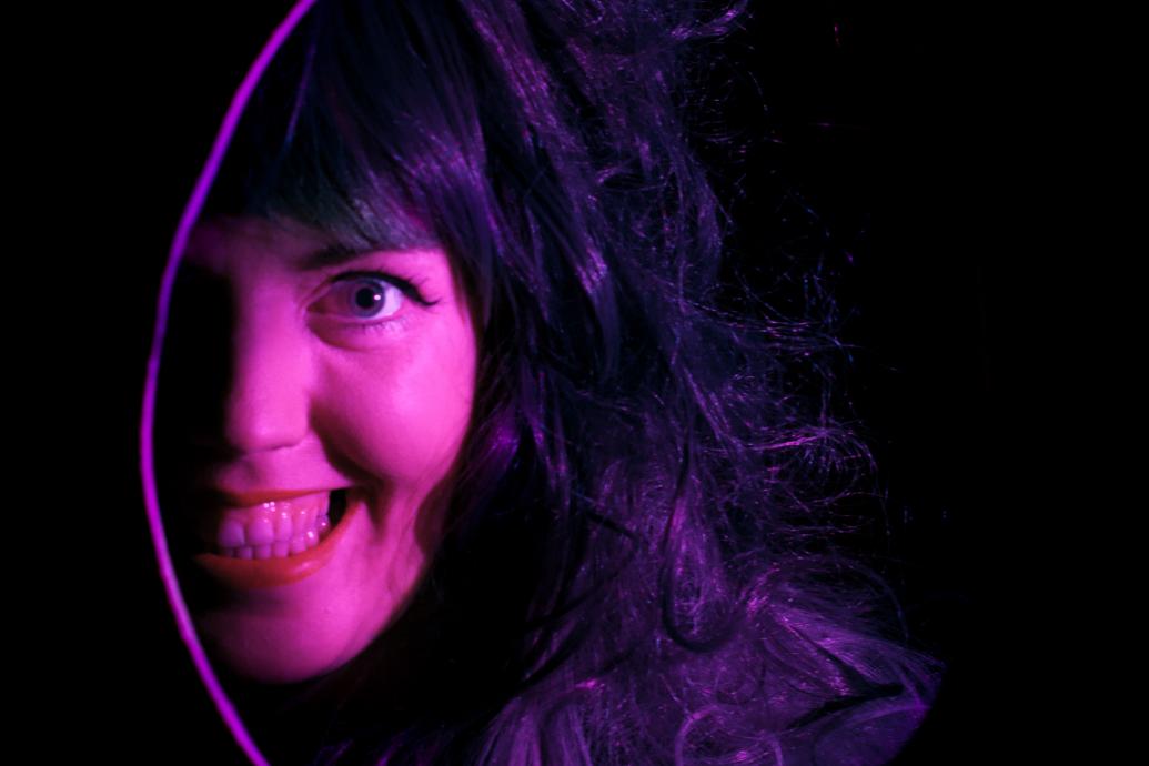 A woman's smiling face, lit in purple, is visible through a oval hole in a black space
