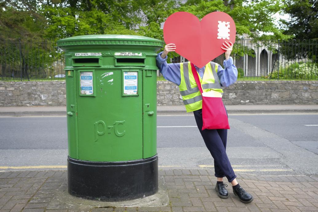 Postwoman holding a heart envelope beside an Irish postbox