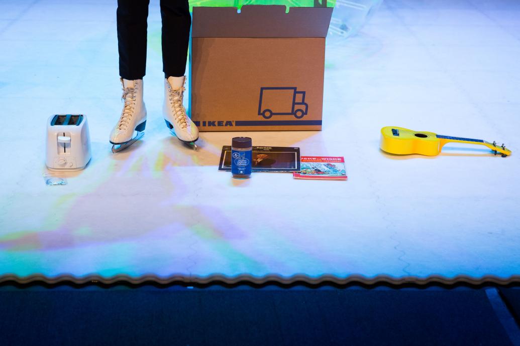 A person stands in ice-skates on an ice rink, only their feet and calves are visible, to the left is a toaster, to the right an Ikea box, a jar of coffee, a record, a book and a ukulele 