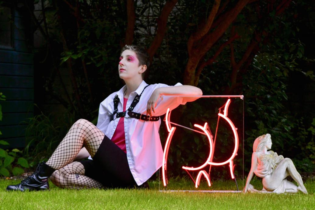 Púcaparty Glitter Hole, A woman sits on the grass leaning against a neon sign in a pose like the White Lady of Cabra statue sitting behind her