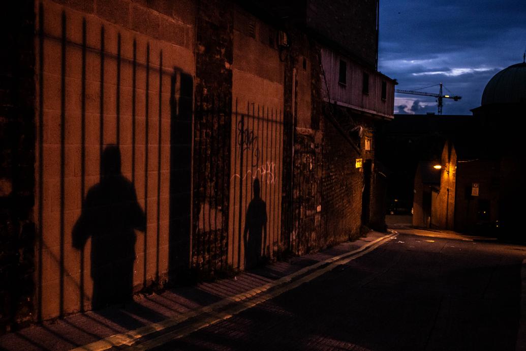 The shadows of two people, standing apart against railings, are projected onto an alley wall at night