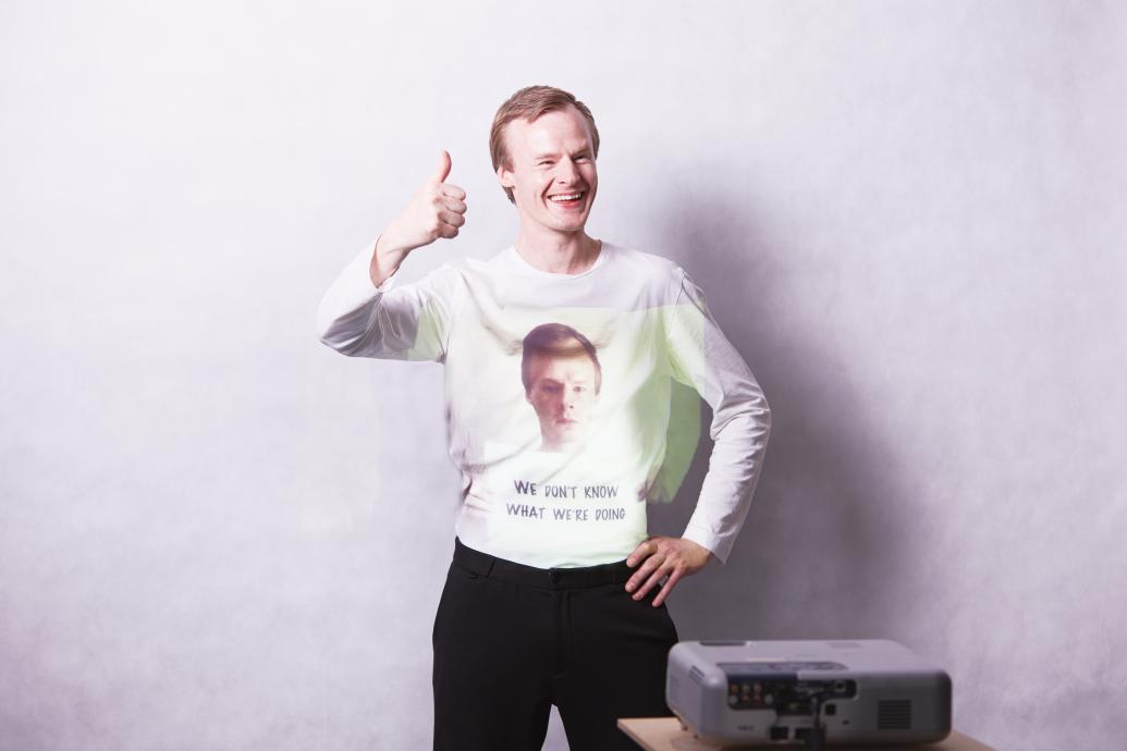 A man smiles, giving a thumbs up, as an image of himself frowning and holding a sign saying "We don't know what we're doing" is projected onto his top