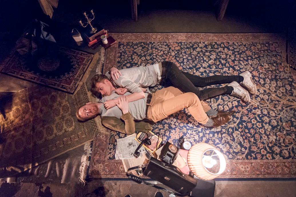 Two men lie together on a carpet holding each other