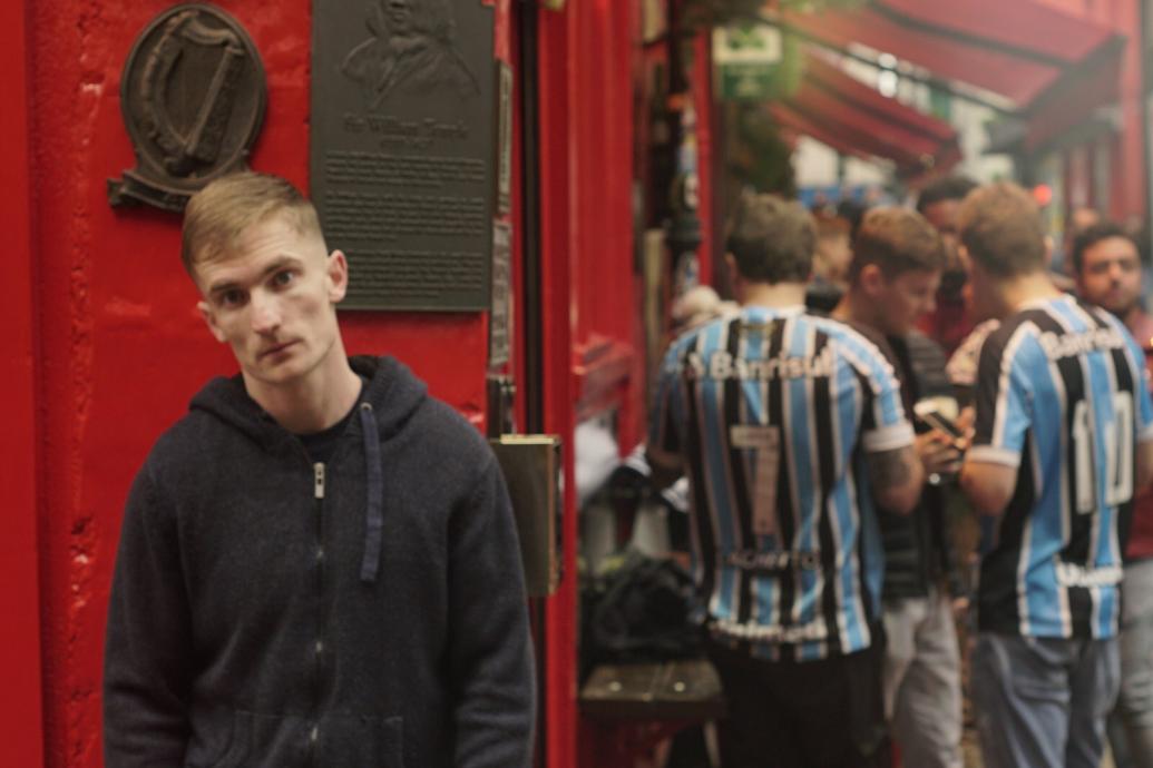 A man stands outside against the wall of a pub facing the camera as a group of men in jerseys linger around the corner from him 