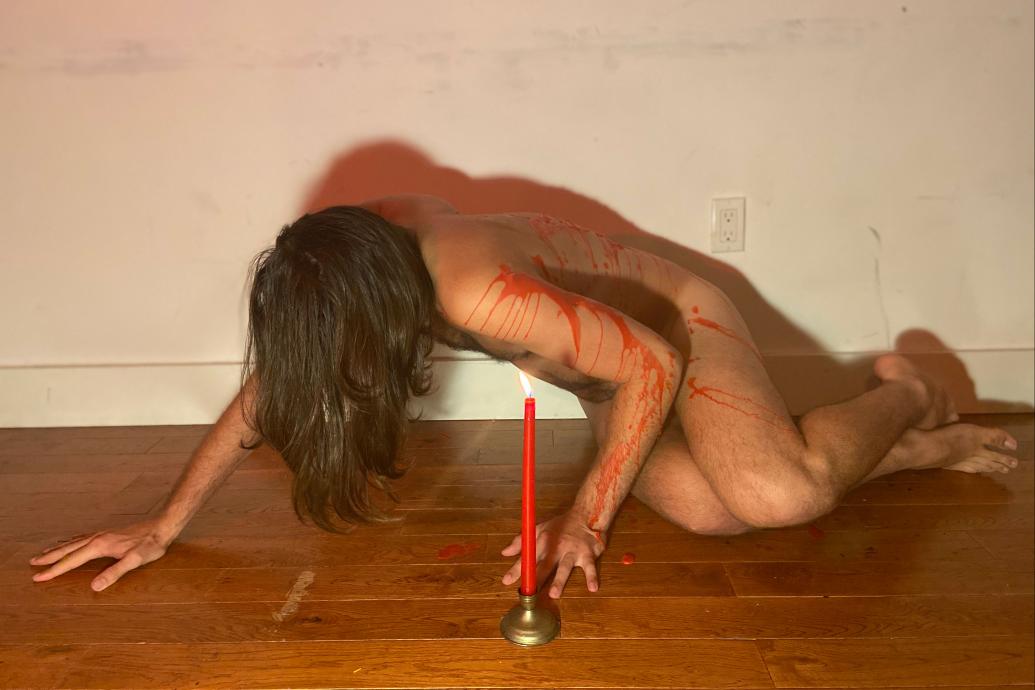 A man crouched over a wooden floor covered in red wax with a red candle in front of him