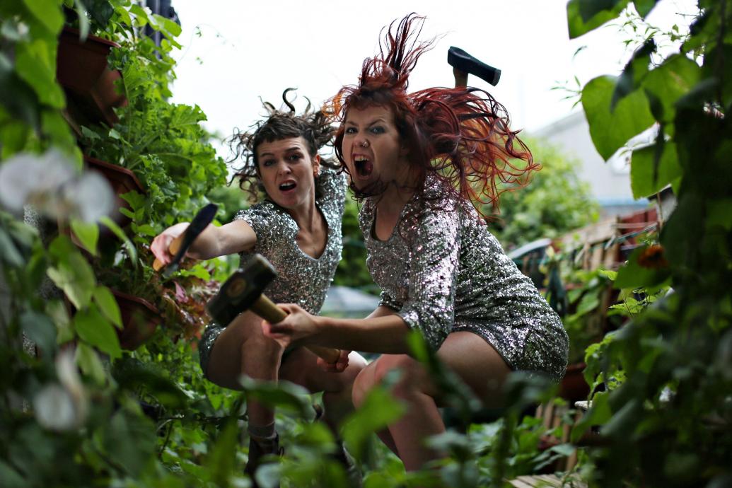 Two fierce women in sequin dresses crouch with garden weapons in leafy surroundings