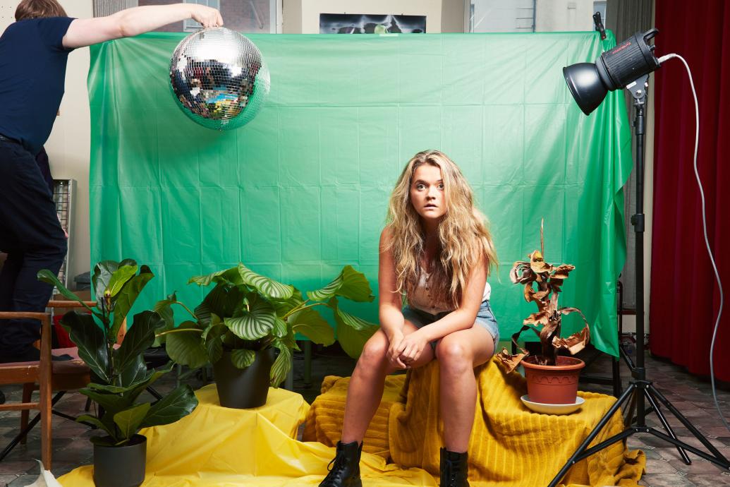 Woman sitting by green screen surrounded by props of plants and a disco ball held in the air by a man
