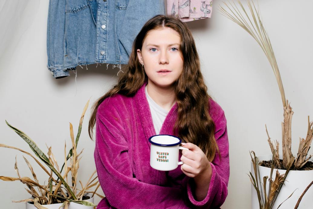 Young woman seated with a cup of tea surrounded by clothes and dead plants