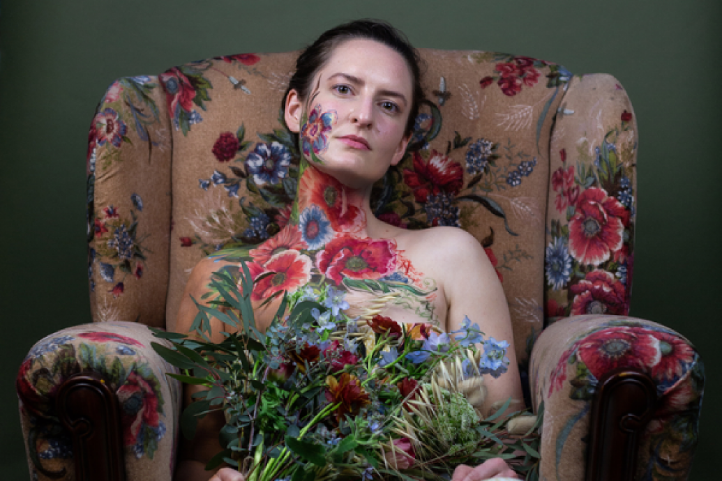 A woman with a floral design painted on her skin with flowers sitting in a floral upholstery armchair