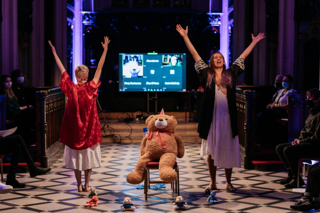 Two performers with hands in the air either side of a large teddy bear in a chapel