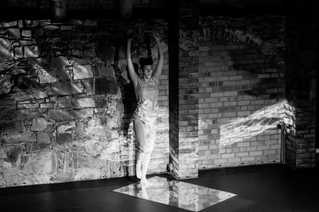 Dancer Isabella Oberlander performing on stage standing against a stone wall on a reflective surface